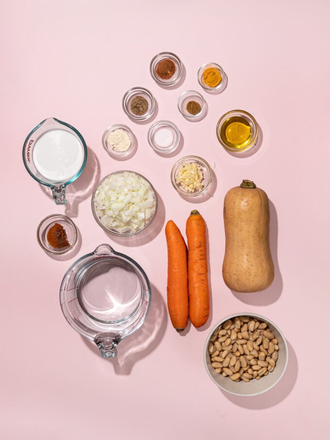 whole butternut squash, 2 carrots, and small bowls of ingredients, arranged on a pink backdrop