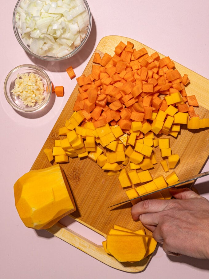 hand dicing butternut squash and carrot on wooden chopping board next to bowls of onion and garlic