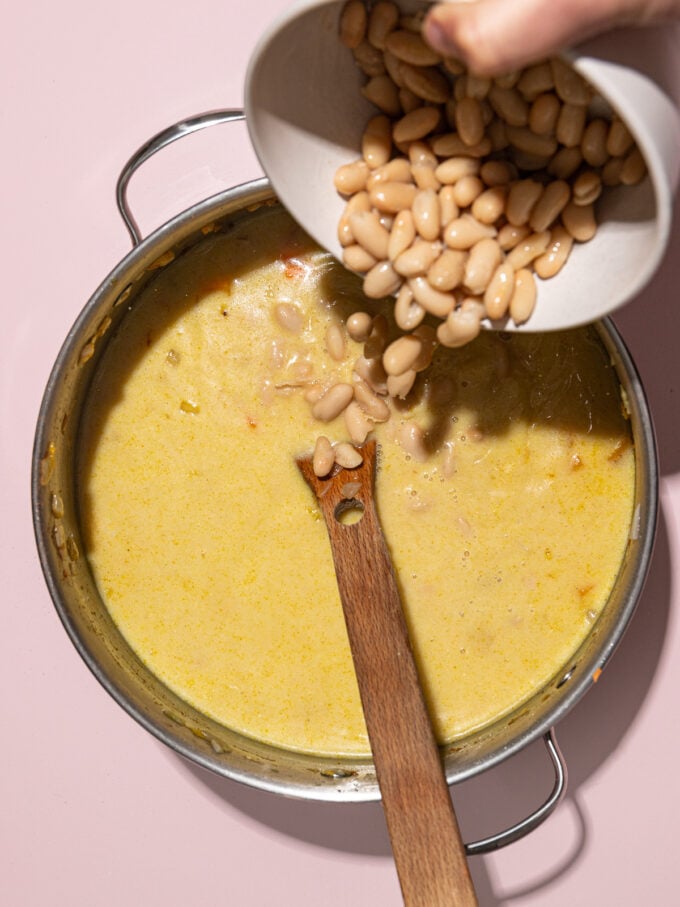 hand pouring white beans out of white bowl into soup pot