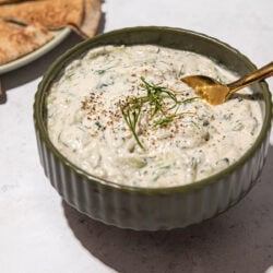 bowl of vegan tzatziki with gold spoon inserted into it. a plate of sliced pita bread beside it.