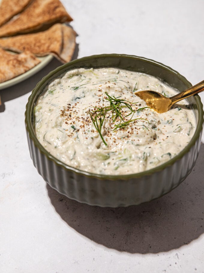 bowl of vegan tzatziki with gold spoon inserted into it. a plate of sliced pita bread beside it.