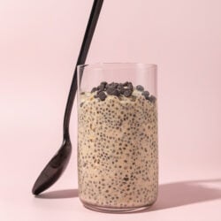 glass filled with cookie dough overnight oats with a black spoon leaned up against it, on a pink background