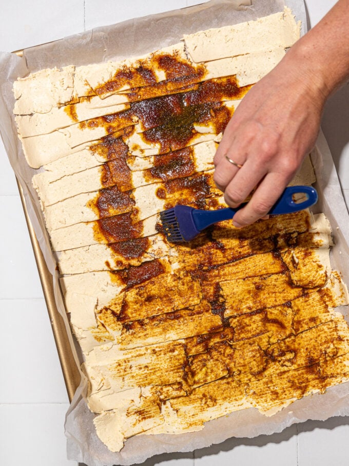 hand brushing shawarma sauce onto shaved tofu on baking sheet